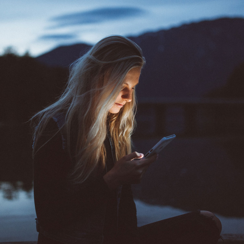 Junge Frau schaut in der Dämmerung am See auf ihr Handy